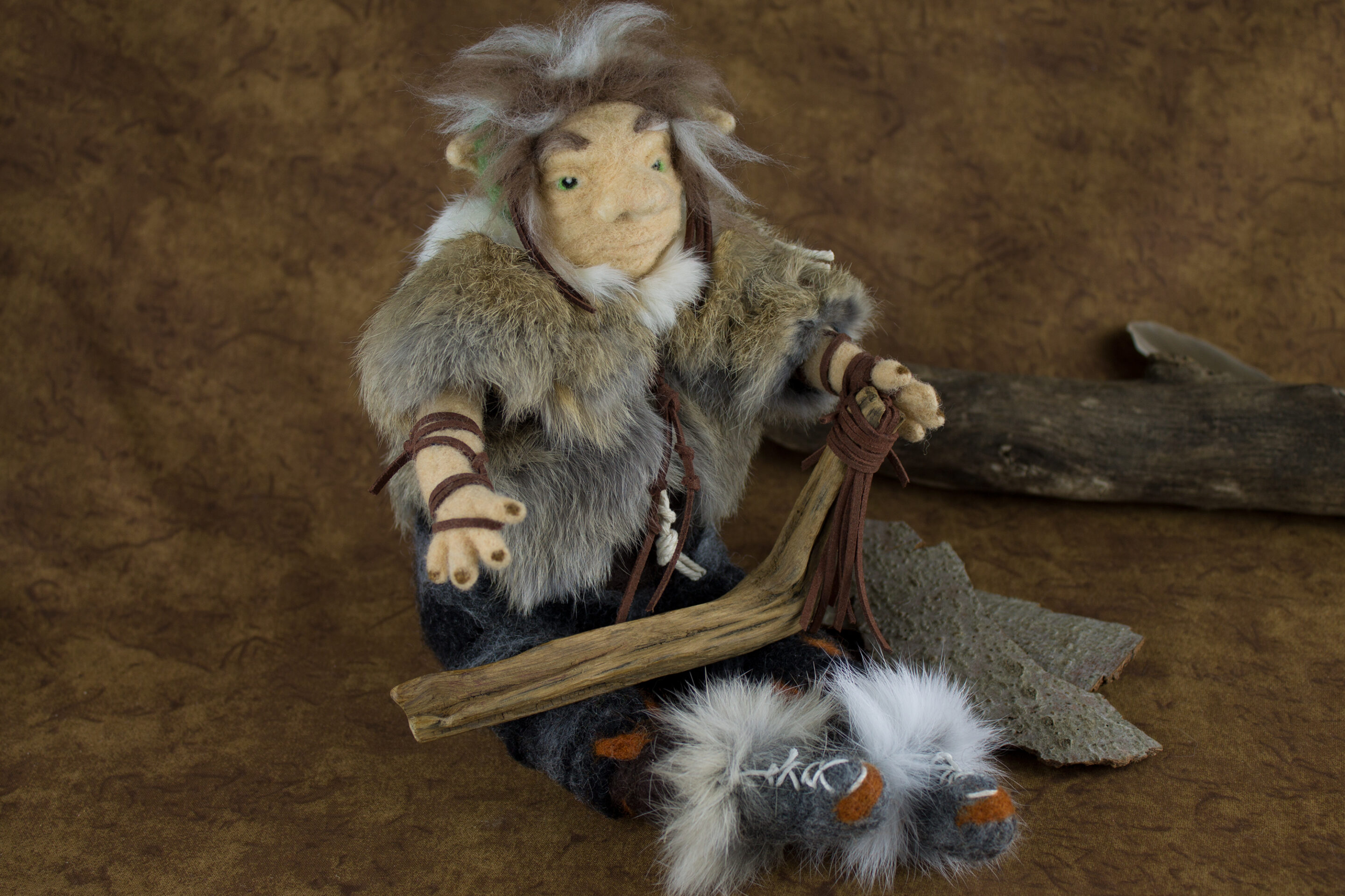 Filzfigur, sitzender Naturgeist mit einer Jacke aus Kaninchenfell, er hält einen Holzstab auf den Beinen