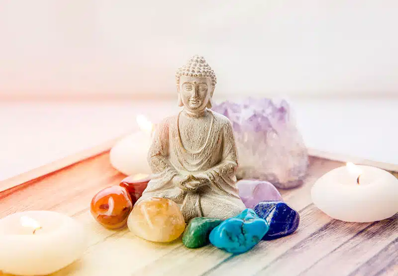 Buddhafigur, klein, weiß, umgeben von bunten Edelsteinen und zwei kleinen, runden Kerze auf einem Tablett