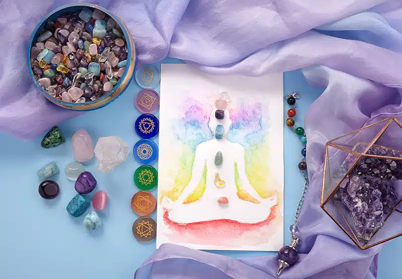 eine Karte mit einer Chakra-Zeichnung, umgeben von Chakrensteinen und einem fliederfarbenen Tuch und anderen Edelsteinen