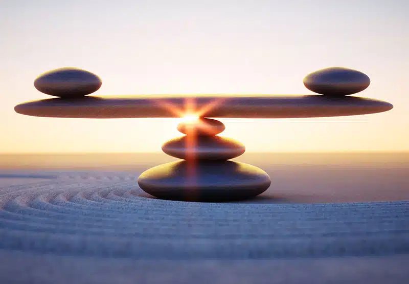 wellness-hypnose-800 Steine an einem Stand, die zu drit zu einem kleinen Turm gelegt sind, auf dem ein großer flacher Stein ruht. Auf diesem ruhen an jedem Ende ein weiterer, flacher Stein als Symbol der Balance.